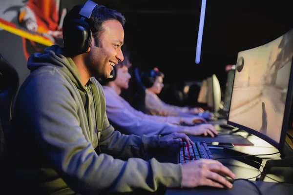 Side view of smiling indian man in headphones playing video game on computer with blurred team in cyber club