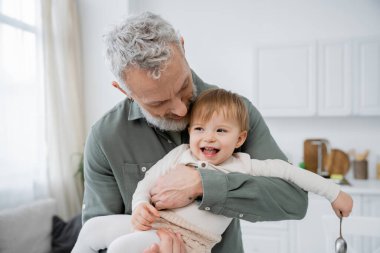 bearded mature man embracing carefree granddaughter holding spoon in kitchen clipart