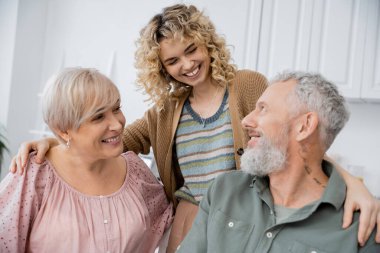 smiling woman with blonde wavy hair embracing joyful middle aged parents in kitchen at home clipart