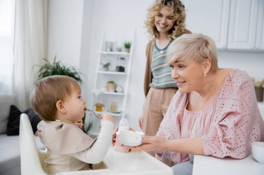 Mutfakta kahvaltıda elinde kaşıkla kucağında bebek torunu taşıyan orta yaşlı bir kadın.