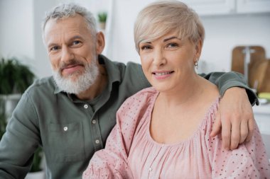 happy middle aged woman and bearded man smiling at camera while sitting in kitchen at home clipart