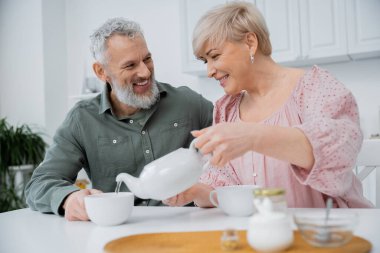 Mutlu orta yaşlı kadın mutfaktaki sakallı kocanın yanına çay koyuyor.