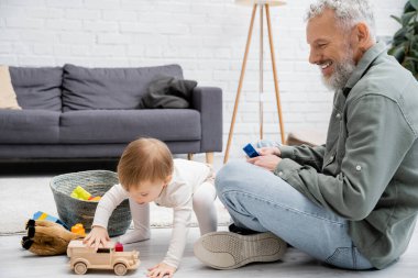 bearded man smiling near toddler granddaughter playing with toy car near building blocks on floor in living room clipart