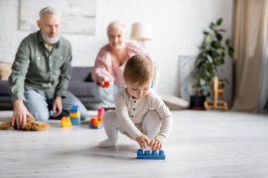 baby girl playing with building blocks on floor in living room near grandparents on blurred background clipart