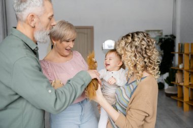 Sakallı bir adam oyuncak atı neşeli bir torunun ve ailenin yanında oturma odasında tutuyor.