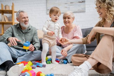 joyful woman holding hand of toddler daughter near happy parents and toys on floor in living room clipart