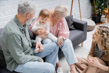 cheerful woman sitting on floor in living room and looking at mature parents playing with granddaughter on couch clipart