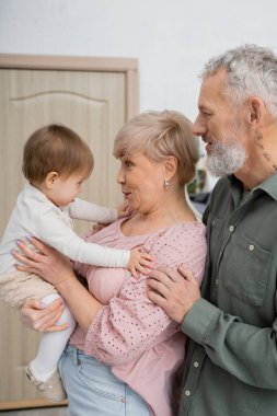 side view of happy woman looking at little granddaughter near bearded husband at home clipart