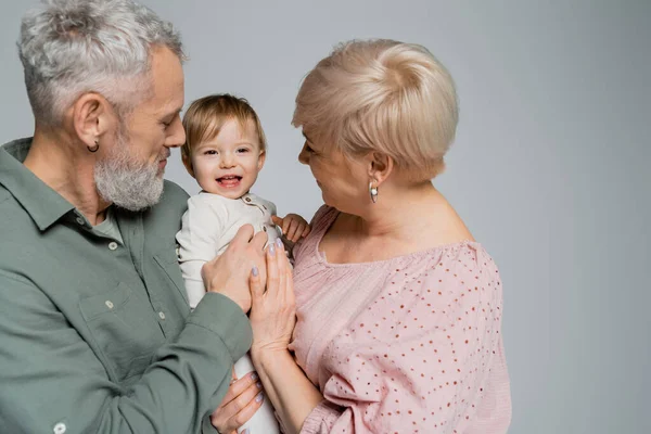 cheerful baby girl smiling near grandmother and bearded grandpa isolated on grey