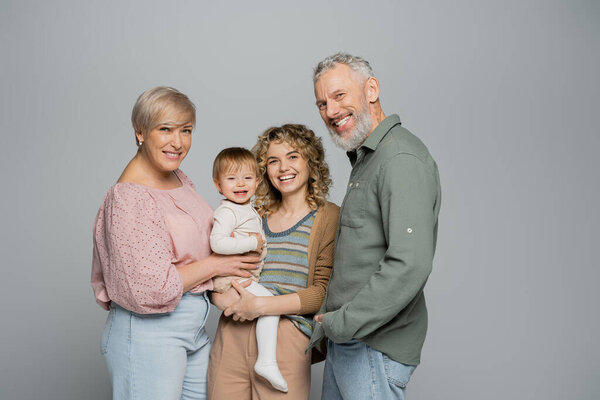 cheerful woman holding toddler daughter near happy senior parents isolated on grey