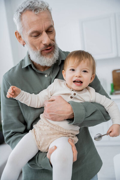 bearded grey haired man holding cheerful daughter with spoon in kitchen