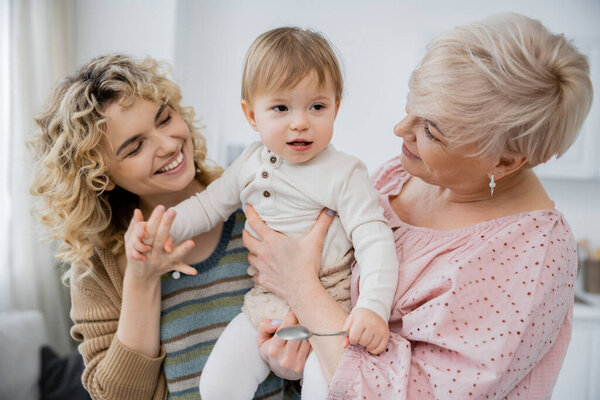 joyful women looking at toddler girl holding spoon in kitchen