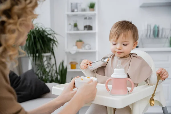 在厨房吃早饭的时候 蹒跚学步的小女孩拿着勺子坐在妈妈旁边的婴儿椅子上 手里拿着碗 — 图库照片