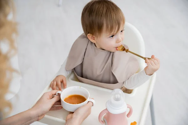 小女孩拿着勺子 坐在靠近妈妈的婴儿椅子上 厨房里拿着碗 一边吃着美味的南瓜 — 图库照片