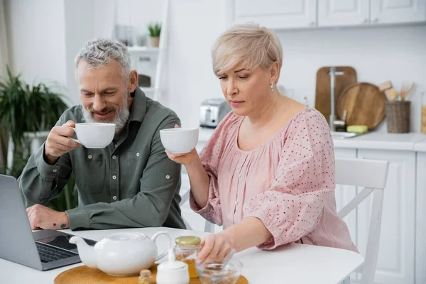 middle aged woman reaching jar of honey while drinking tea with happy husband near laptop in kitchen