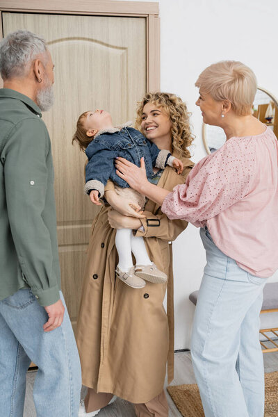 cheerful woman in trench coat holding little daughter in denim jacket while visiting mature parents at home