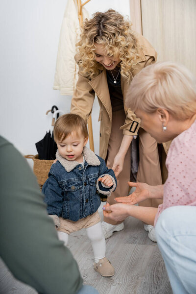 cheerful woman in trench coat and little child in denim jacket near blurred grandparents in hall at home