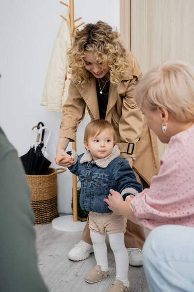 smiling woman in trench coat and toddler girl in denim jacket visiting grandparents at home
