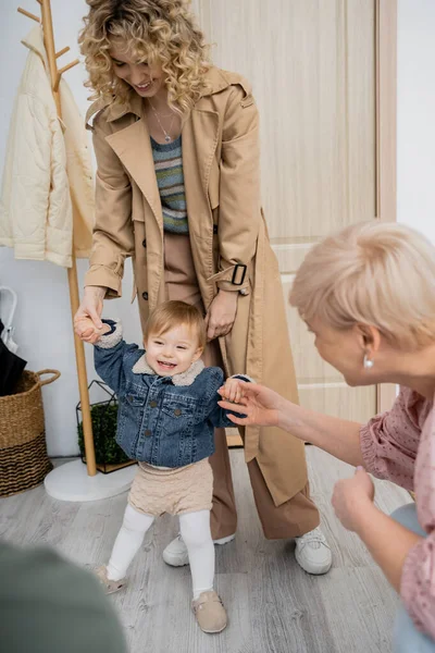 在家里 穿着牛仔夹克的蹒跚学步的小女孩在妈妈身边微笑 奶奶在大厅里笑 — 图库照片