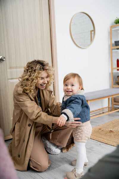 curly woman in trench coat hugging carefree and cheerful daughter in denim jacket near door at home