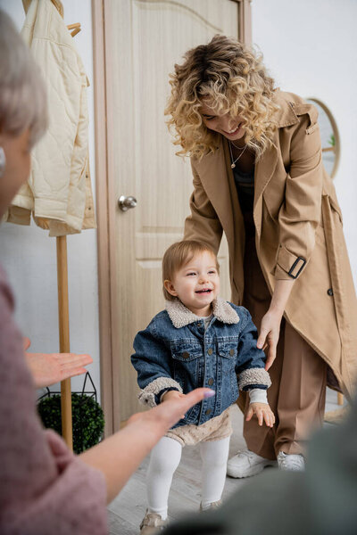blurred woman meeting daughter with overjoyed child in denim jacket near door at home