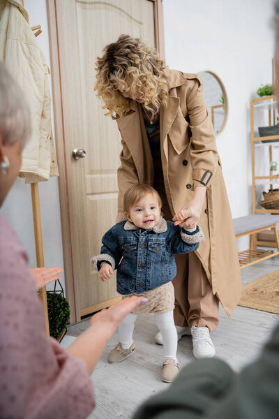 woman in trench coat and little girl in denim jacket visiting grandparents at home on blurred foreground