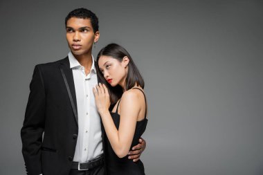 Asyalı seksi bir kadın siyah ceketli, zarif bir Afro-Amerikan erkeğine yaslanıyor. Gri üzerine izole edilmiş.