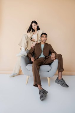 full length of interracial couple in stylish pantsuits posing near armchair on beige background clipart