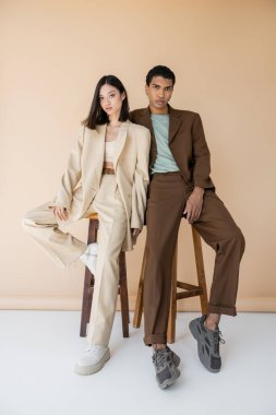 full length of multiethnic couple in stylish pantsuits posing near stools on beige background clipart