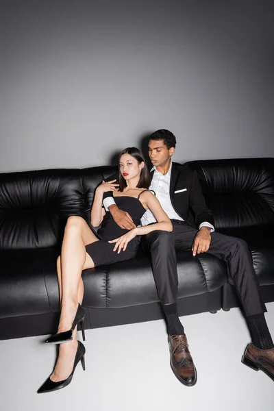 全长的不同种族的年轻夫妇穿着雅致的服装 坐在灰色背景的黑色皮沙发上 — 图库照片