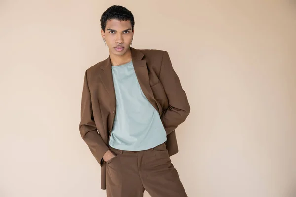 young african american man in brown suit standing with hand in pocket and looking at camera isolated on beige