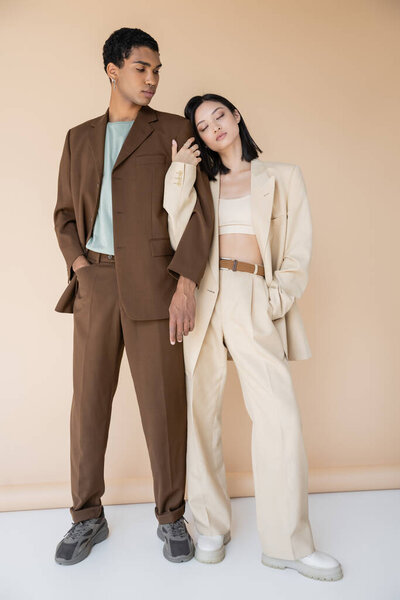 full length of stylish asian woman with closed eyes leaning on african american man in brown suit on beige background