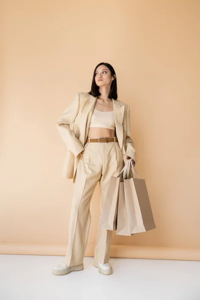 アジアの女性の完全な長さでショッピングバッグポージングでスタイリッシュな象牙のパンツでベージュの背景 — ストック写真