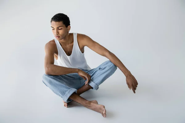 barefoot african american man in jeans sitting with crossed legs on grey background