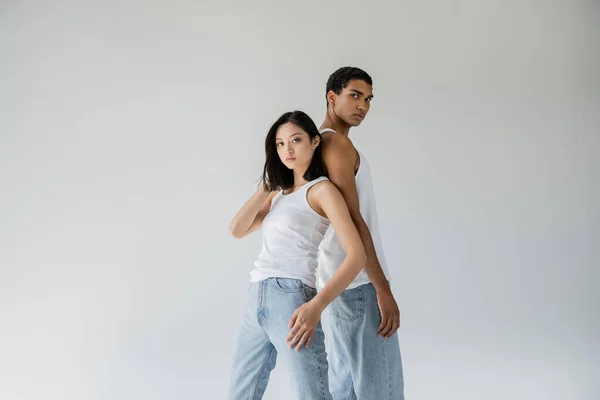 青のジーンズと白のタンクトップを着た若いスタイリッシュな異人種間のカップルがグレーで隔離されたカメラを — ストック写真