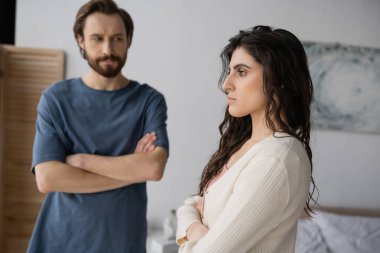 Offended brunette woman crossing arms near blurred boyfriend in bedroom  clipart