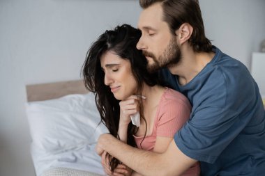 Sakallı adam yatak odasında peçeteyle tatminsiz kız arkadaşına sarılıyor. 