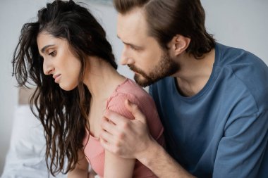 Caring bearded man calming down displeased girlfriend in bedroom at home  clipart