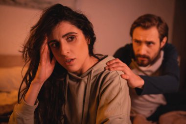 Sad woman touching head near blurred boyfriend at home in evening  clipart