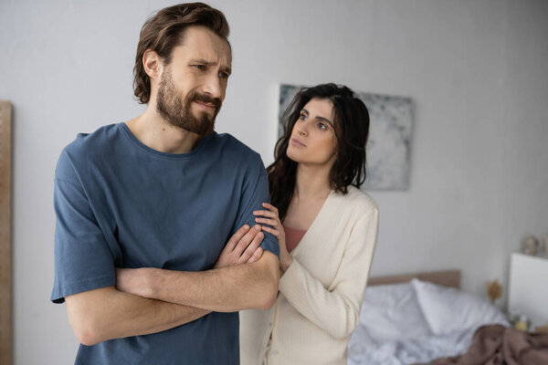 Недовольный мужчина скрещивает руки рядом с расплывчатой брюнеткой в спальне 