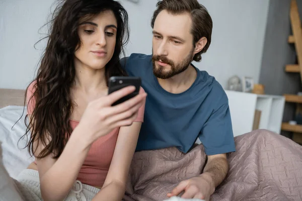 ブルネット女性でPajama使用スマートフォン近くに深刻なボーイフレンドオンベッド — ストック写真