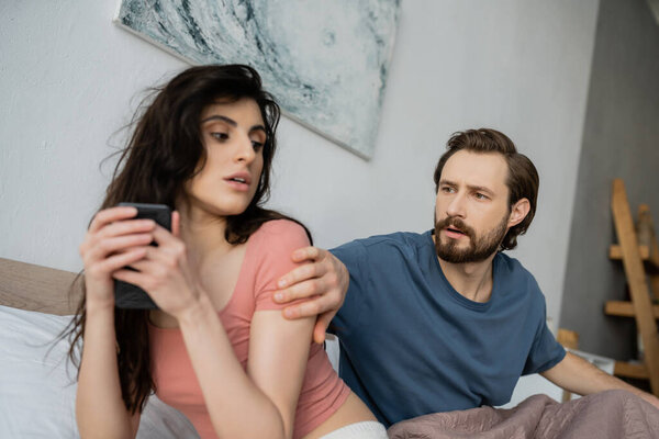 Jealous man looking at brunette girlfriend hiding smartphone in bedroom at home 