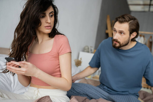 Brunette woman hiding smartphone near blurred angry boyfriend on bed 