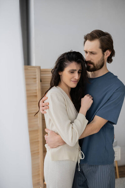 Bearded man in pajama hugging dissatisfied girlfriend at home 