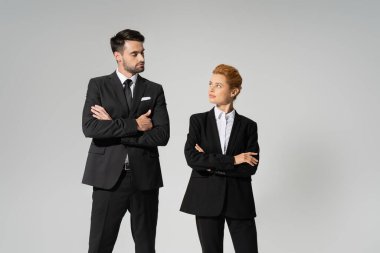 skeptical business people in black pantsuits standing with folded arms and looking at each other isolated on grey clipart