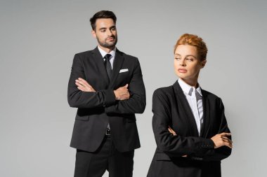 skeptical businessman in stylish suit looking at redhead woman in formal wear isolated on grey clipart
