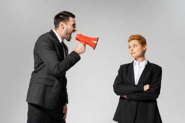 Öfkeli patron megafonla bağırıyor, kayıtsız kızıl saçlı çalışanın yanında, kollarını kavuşturmuş gri renkte.