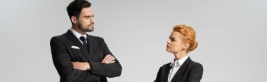 serious and skeptical business people looking at each other while standing with crossed arms isolated on grey, banner clipart