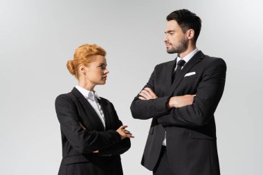 confident businessman looking at serious redhead businesswoman isolated on grey clipart