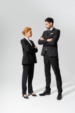 full length of skeptical business people in black suits looking at each other while standing with crossed arms on grey background clipart